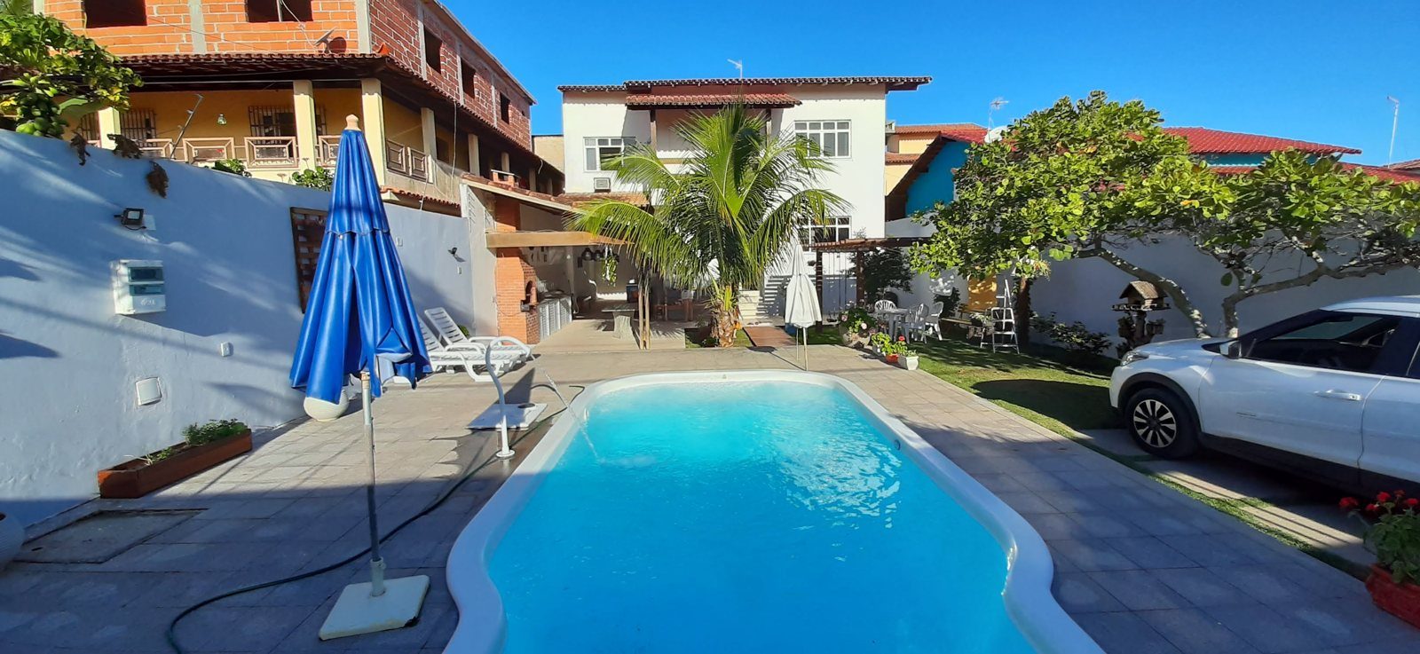 Piscina com a casa ao fundo, coqueiro, espreguiçadeiras e área de lazer